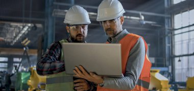 Güvenlik üniformalı iki profesyonel teknisyen ve kasklar imalat fabrikasında dizüstü bilgisayarla çalışıyorlar. Olgun mühendis ve yetişkin işçi konuşuyor ve depoda tartışıyorlar..