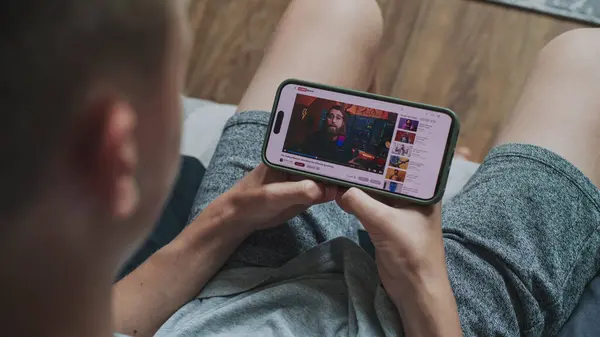 stock image Caucasian boy watches educational video of blogger about stock trading using mobile phone while sitting on sofa. Smartphone screen shows video hosting website interface with video content. Close Up.