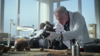 Kıdemli arkeolog modern arkeoloji laboratuvarında tarih öncesi insan kafatası ve kemiklerinin fosil kalıntılarının fotoğrafını çekiyor. Bilim adamları arka planda çalışıyor. Arkeoloji ve paleoantropoloji.