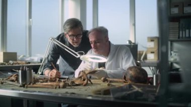 Kıdemli paleoantropolog tarih öncesi insan kalıntılarını arkeoloji laboratuvarında araştırıyor, profesyonel araçlar kullanıyor, iş arkadaşlarıyla konuşuyor. Erkek arkeolog süreci izler, not defterine notlar alır..