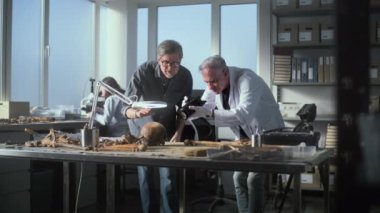 Kıdemli paleoantropolog, tarih öncesi insan kafatasının fosil kalıntılarını ve kamera kullanarak arkeoloji laboratuvarındaki meslektaşlarıyla birlikte kemiklerin fotoğrafını çekiyor. Kadın bilim adamı arka planda çalışıyor..