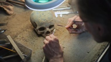 Yetişkin bir arkeoloğun nesli tükenmiş antik bir insanın kafatasını profesyonel aletlerle lamba altından temizlemesine yakın çekim. Erkek bilim adamı, arkeoloji laboratuvarında çalışan fosil kalıntılarını inceliyor..
