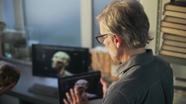 Arkeoloji laboratuvarı: İki bilim adamı fosil kafatası üzerinde çalışıyor, dijital tablet ve kişisel bilgisayar ekranında gösterilen soyu tükenmiş antik insan kafasının üç boyutlu modelleme görselleştirmesini tartışıyor.