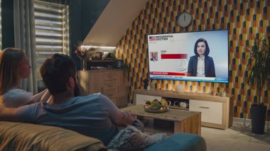 ZAPORIZHYA, UKRAINE - 7 Haziran 2024: Beyaz çift, ABD başkanlık seçimlerinin sonuçlarının koltukta oturduğu televizyon haberlerini izliyor. Donald Trump seçimi kazandı. Kadın televizyon haberleri..