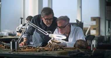 Kıdemli paleoantropolog, tarih öncesi insan kalıntılarını arkeoloji laboratuvarında inceliyor, profesyonel araçlar kullanıyor, meslektaşıyla objeyi tartışıyor. Bilim adamı not defterine notlar alır..