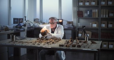 Modern arkeoloji laboratuvarı: Kıdemli arkeoloji, profesyonel araçlarla lambanın altındaki fosil kafatasını temizliyor, soyu tükenmiş antik bir insanın iskelet kalıntılarını inceliyor. Erkek meslektaşım arka planda çalışıyor..