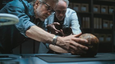 İki arkeolog konuşuyor ve tripodun üzerinde profesyonel kamera kullanarak tarih öncesi insan kafatasının fosil kalıntılarının fotoğrafını çekiyor. Bilim adamları modern arkeoloji laboratuvarında çalışıyorlar. Arkeoloji kavramı.