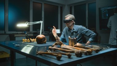 Geceleyin arkeoloji laboratuvarı: VR kulaklıktaki olgun arkeolog artırılmış gerçeklik teknolojisini kullanarak fosil kafatasını inceliyor. Tablet bilgisayar ve iskelet kalıntıları masada soyu tükenmiş eski bir insana ait..