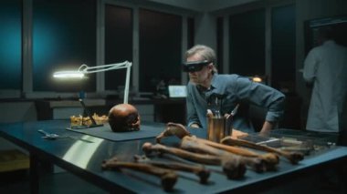 Geceleyin arkeoloji laboratuvarı: VR kulaklıktaki olgun arkeolog artırılmış gerçeklik teknolojisini kullanarak fosil kafatasını inceliyor. Tablet bilgisayar ve iskelet kalıntıları masada soyu tükenmiş eski bir insana ait..