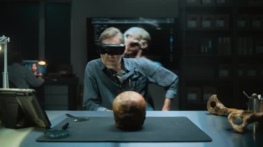 VR kulaklığındaki olgun arkeolog, artırılmış gerçeklik kullanılarak gece vakti arkeoloji laboratuvarında soyu tükenmiş antik bir insanın fosil kafatasını inceliyor. Arka planda 3 boyutlu kafa modeli olan büyük ekran.