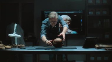 Olgun arkeolog, tripodun üzerinde kamera kullanarak tarih öncesi insan kafatasının arkeoloji laboratuvarında fosil kalıntılarının fotoğrafını çekiyor. İş arkadaşı ve büyük ekran. Arka planda 3 boyutlu kafa modeli var..