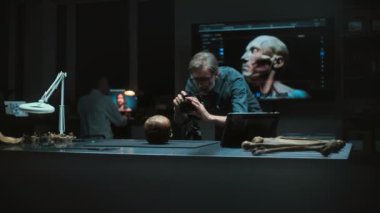 Olgun arkeolog, tripodun üzerinde profesyonel kamerayla antik insan fosil kafatasını arkeoloji laboratuvarında fotoğraflıyor. Arka planda kafanın 3 boyutlu görselleştirilmiş iş arkadaşı ve büyük ekran.