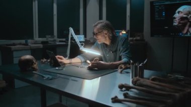 Arkeolog ölçümler yapıyor, lambanın altındaki fosil kemikleri inceliyor, tablet bilgisayar kullanıyor. Bilim adamları geceleri arkeoloji laboratuvarında çalışıyorlar. Arka planda 3 boyutlu insan modeli olan büyük ekran.