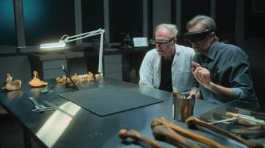 VR kulaklık takan iki arkeolog, artırılmış gerçeklik ve tablet bilgisayar kullanarak kemiklerle ilgili tarihi bir araştırma yürütüyor. Bilim adamları geceleri arkeoloji laboratuvarında çalışıyorlar..