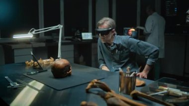 Geceleyin arkeoloji laboratuvarı: AR 'da olgun bir arkeolog sanal gerçekliği kullanarak tarih öncesi soyu tükenmiş insanların kafatası kalıntıları üzerinde çalışıyor. Arkaplanda 3 boyutlu görselleştirmesi olan bilim adamı ve büyük ekran.