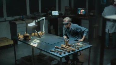Geceleyin arkeoloji laboratuvarı: VR kulaklıklı olgun bir arkeologun iskelet kalıntıları üzerine bilimsel araştırma yürüterek gelişmiş gerçekliği kullanması. İş arkadaşı arka planda çalışır.