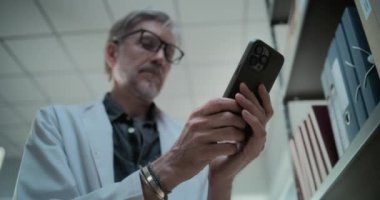 Laboratuvar önlüğü giymiş yetişkin bir bilim adamının sohbet ederken, internette veya sosyal ağda dizinlerle rafların yanında dururken cep telefonu kullanırken yakın plan çekimleri. Modern arkeoloji laboratuvarı. Düşük Açı.