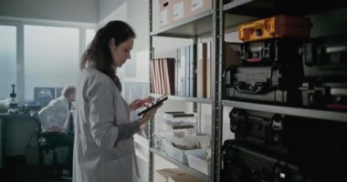 Modern arkeoloji laboratuvarı: Laboratuvar önlüklü kadın bilim adamı tablet bilgisayarla rafa geliyor, bilimsel veya tarihi araştırmalar için klasör alıyor. Erkek arkadaş arka planda bilgisayar üzerinde çalışıyor..