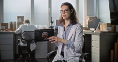 Kadın laboratuvar çalışanı veya bilim adamı kameraya konuşuyor, arkeoloji laboratuvarında popüler bilim filmi için yapılan röportaj sırasında tablet bilgisayarda insan kafasının üç boyutlu görüntüsünü gösteriyor. Sabit Çekim.