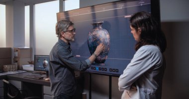 Erkek ve kadın bilim adamları veya arkeologlar kültürel eserleri tartışıyor ve inceliyor, gelişmiş arkeoloji laboratuvarında tarihi araştırmalar yapıyorlar. Büyük ekranda antik kil vazonun 3D görüntüsü gösteriliyor.