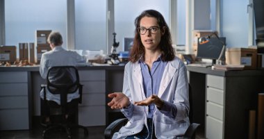 Laboratuvar önlüklü genç kadın bilim adamı, arkeoloji laboratuvarında otururken kameraya konuşuyor ve popüler bilim filmi için röportaj veriyor. Erkek meslektaşım arka planda çalışıyor. Sabit Çekim.