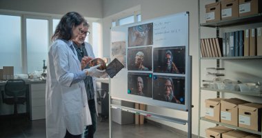 Erkek ve kadın bilim adamları tablet bilgisayar kullanarak antik bir adamın fosil kafatasını inceliyorlar. Arkeologlar arkeoloji laboratuvarında çalışırlar. İnsan kafası anatomisinin 3 boyutlu görselleştirilmiş resimlerini içeren tahta.
