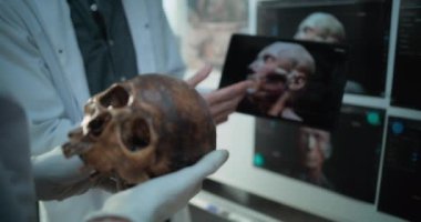 Bilim insanları fosil kafatası anatomisini tarih öncesi insan kafasının 3 boyutlu modelini kullanan tablet bilgisayarla araştırıyor, arkeoloji laboratuvarında çalışıyorlar. Arka planda antik insanların yazılı resimlerinin olduğu tahta.