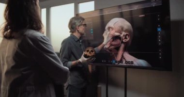 Erkek ve kadın bilim adamları veya arkeologlar konuşurlar, fosil kafatası üzerinde çalışırlar ve gelişmiş arkeoloji laboratuvarlarında tarihi araştırmalar yaparlar. Büyük ekranda eski insan kafası anatomisinin 3 boyutlu görüntüsü var..