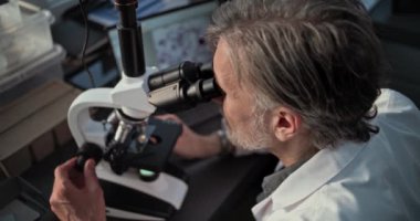 Olgun mikrobiyolog mikroskop altında fosil analizi yapar, laptop ekranındaki mikroorganizmalara bakar. Bilim adamı, arkeoloji laboratuvarında gelişmiş ekipmanlarla çalışıyor. Yüksek Açı.