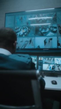 Afrikalı Amerikalı erkek gözetleme operatörü modern izleme merkezinde bilgisayardaki güvenlik kameralarıyla çalışıyor. Çoklu monitörlerdeki CCTV görüntüleri ve büyük dijital ekran. Arkadan bak. Dikey çekim.