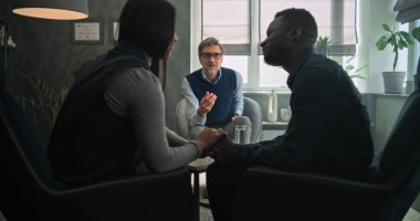 Başarılı çiftler terapisinden sonra sandalyede oturan ve el ele tutuşan çok ırklı çiftin uzlaşması. Olgun psikoterapist genç aileyle konuşur, danışmanlık seansı düzenler. Psikoloji.