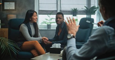 Mutlu çok ırklı çiftler el ele otururlar, gülerler, birbirlerine bakarlar, çiftler terapisinde kavga ettikten sonra uzlaşırlar. Psikologla başarılı bir danışmanlık seansı yapan hassas genç aile..