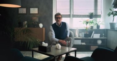 Profesyonel bir terapist ya da gözlüklü bir psikolog kameraya bakıp şık bir psikoloji ofisinde oturuyor. Grup terapisi, danışmanlık seansı. Ruh Sağlığı.