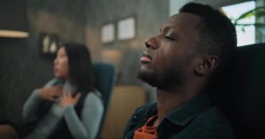 Çift terapisi sırasında kız arkadaşını dinleyen Afrikalı Amerikalı bir adama yakın çekim. Arka planda, odaklanmamış Asyalı kadın terapistle terapi seansında ilişki sorununu tartışıyor..