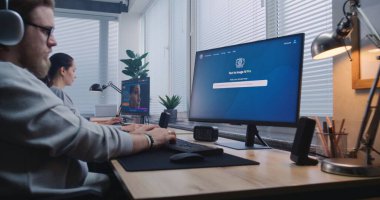 Kulaklıklı yetişkin adam bilgisayardaki yapay zeka görüntü jeneratörüne giriyor. Yapay zeka sohbet robotu, bilim kurgu robotunun detaylı bir portresini oluşturuyor. Bayan meslektaşım arka planda bilgisayar üzerinde çalışıyor..