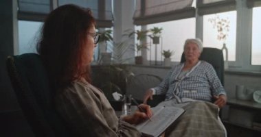 Psikoterapi ofisinde kadın uzman ve sandalyelerde oturan yaşlı kadın hasta. Yaşlı kadın, doktorların endişe ile ilgili sorularını cevaplarken gergin. Terapist cevapları yazıyor..