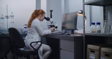 Kadın mikrobiyolog mikroskop altında inceleniyor. Profesyonel bilim adamı yüksek teknolojili ekipman kullanarak örnek analizi yapar. Bilgisayar ekranında hücrelerin veya bakterilerin görüntüsü var. Mikrobiyoloji.