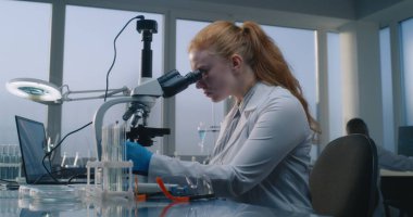 Parlak tıp bilimi laboratuvarı: Profesyonel mikrobiyolog dizüstü bilgisayar kullanır, mikroskop altında test örneklerini inceler. Kadın bilim adamı, biyokimyacı yüksek teknolojili ekipmanlarla çalışıyor, uyuşturucu geliştiriyor..