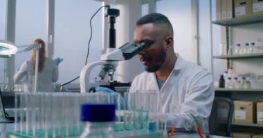 Parlak tıp bilimi laboratuvarı: Afrikalı Amerikalı mikrobiyolog mikroskop altında test örneklerini analiz eder, ilaçlar ve aşılar geliştirir. Bilim adamı ve kimyager yüksek teknoloji ekipmanları ve dizüstü bilgisayarla çalışıyor..