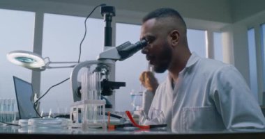 Beyaz önlüklü Afrikalı Amerikalı erkek mikrobiyolog veya kimyager aşı geliştirmek için mikroskop altında yeni virüs örneklerini inceliyor. Arka planda çalışan kadın bilim adamı. Tıp ve mikrobiyoloji.