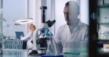 Afrikalı Amerikalı bilim adamı mikroskop altında modern tıp laboratuarında yeni ilaç formülleri üzerinde çalışıyor. Arka planda, dişi mikrobiyolog verileri bilgisayara giriyor. Tıp konsepti.