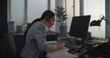 Modern ofiste: Kulaklık takan kadın finans tüccarı bilgisayar başında çalışır, nöbetçi müşteriyle konuşur, hisse senetlerini izler ve hisse senedi piyasasını izler, notlar alır. Uluslararası borsa şirketi.