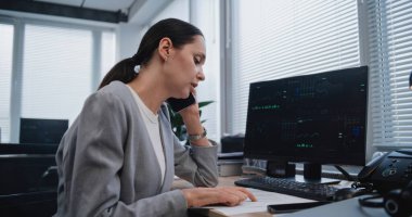 Kadın finans analisti, iş arkadaşıyla cep telefonu kullanarak proje stratejisini tartışıyor. Pazarlama bölümünden uzman, PC monitöründe gösterilen parametrelerle belgelerdeki verileri kontrol ediyor.