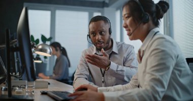 Çağrı merkezi ofisi: Kulaklıktaki iki çoklu etnik yardım hattı operatörü işi tartışıyor, bilgisayarla görüşme yapıyor. Çevrimiçi müşteri desteğinde çalışan teknik destek uzmanları.