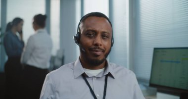 Çağrı merkezi: Afrika kökenli Amerikalı dost canlısı teknik müşteri destek uzmanının kamera karşısında gülümseyen portresi. Arka planda, yardım hattı operatörlerinden oluşan odaklanmamış bir ekip.