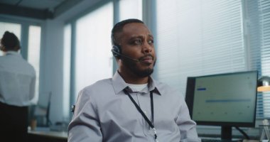 Çağrı merkezi: Afrika kökenli Amerikalı yardım masası uzmanı, gülümseyen, kameraya bakan arkadaş canlısı portresi. Arka planda teknik müşteri destek operatörlerinden oluşan odaklanmamış bir ekip.
