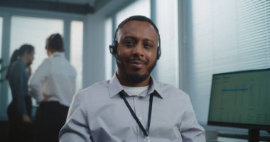 Çağrı merkezi: Afrika kökenli Amerikalı yardım masası uzmanı, gülümseyen, kameraya bakan arkadaş canlısı portresi. Arka planda teknik müşteri destek operatörlerinden oluşan odaklanmamış bir ekip.