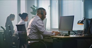 Modern çağrı merkezinde çalışan çok ırklı teknik destek operatörleri ekibi. Afrika kökenli Amerikalı yardım masası uzmanı kulaklıkla konuşuyor, bilgisayar kullanıyor, müşteri hizmetleri sağlayan kameralara bakıyor..