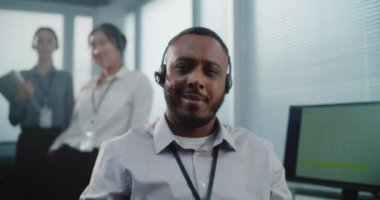 Çağrı merkezi: Afrika kökenli Amerikalı teknik müşteri destek operatörünün kamera karşısında gülümseyen portresi. Arka planda duran yardım masası uzmanları..