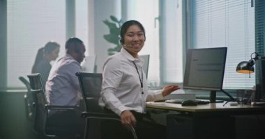 Modern çağrı merkezinde çalışan çok ırklı teknik destek operatörleri ekibi. Asyalı kadın yardım masası uzmanı kulaklıkla konuşuyor, bilgisayar kullanıyor, müşteri hizmetleri sağlayan kameralara bakıyor..